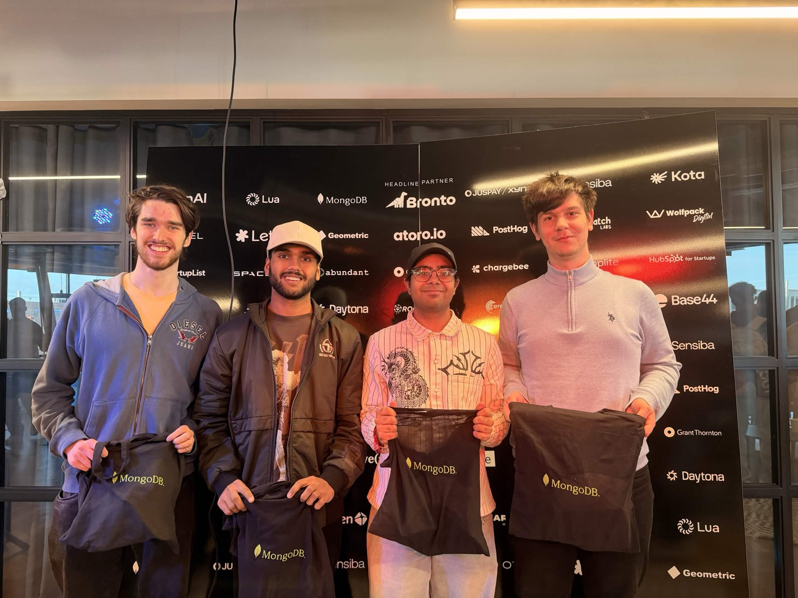 Chirag with three others holding MongoDB tote bags in front of a sponsor wall